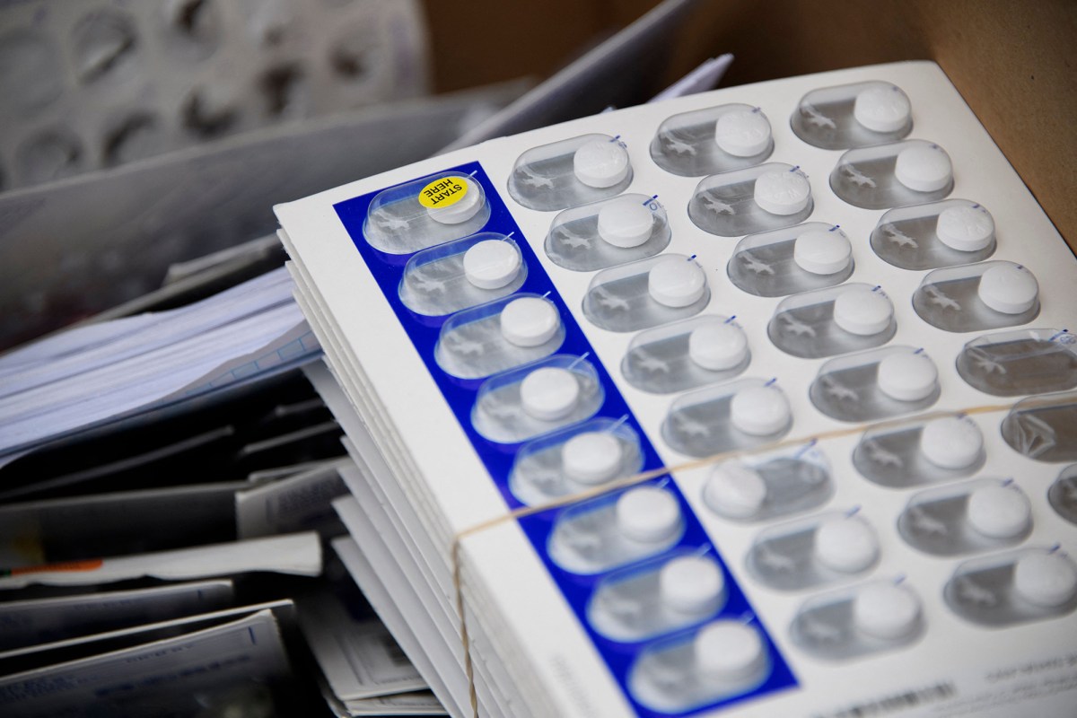 A stack of small white plastic containers with white pills inside plastic bubbles with one with a yellow sticker saying "Start Here". The stack is held with a rubber band and sits on a desk on a stack of papers.