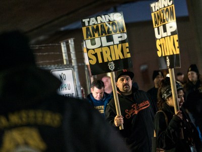 People in sweaters and warm clothing walk with picket signs on signs outside the fence of an Amazon Warehouse. The signs the people are holding say, “ TANNC Amazon ULP Strike.”