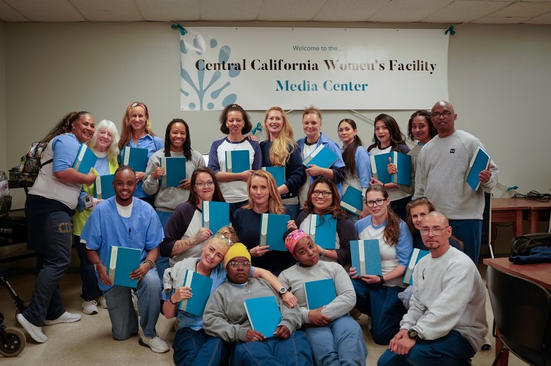 A group of people poses for a photo inside a room with a banner that reads "Central California Women's Facility Media Center." Most individuals are holding blue notebooks and are smiling at the camera. The group includes individuals wearing a mix of casual and uniform-style clothing, standing and seated closely together, reflecting a sense of camaraderie and celebration.