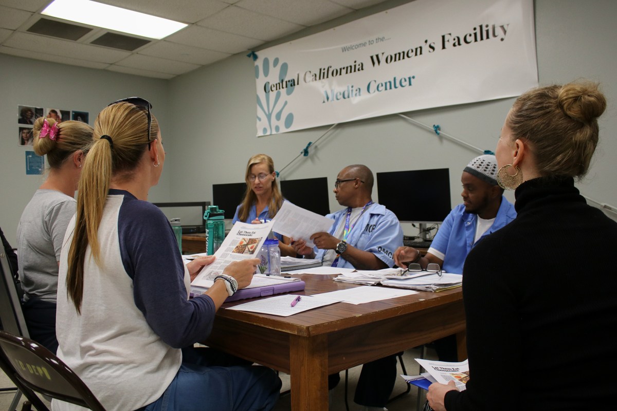 A small group of individuals sits around a table in a classroom-like environment, engaged in discussion and reviewing printed materials. A banner on the wall reads "Central California Women's Facility Media Center." Behind the participants, computer monitors are arranged on desks. The atmosphere appears collaborative and focused, with participants exchanging ideas and referencing documents.