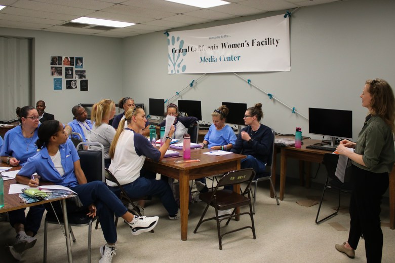 A group of people sits around tables in a classroom-like setting, engaging in discussion and taking notes. A person stands at the front of the room, holding papers, as they lead the session. Behind them, a banner reads "Central California Women's Facility Media Center." Computer monitors are visible on desks along the back wall, and the atmosphere appears focused and collaborative.