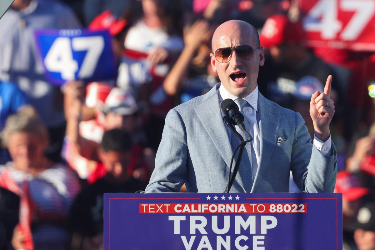 A person in a gray colored suit at a podium labeled “ TEXT CALIFORNIA TO 8802 TRUMP VANCE” talking into a microphone on a stage. Behind them is a crowd of people, with one person holding a blue sign with the number 47 on it.