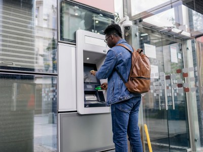 A person with a denim jacket and a backpack uses their credit card at an ATM to withdraw cash. The ATM is outside the bank entrance on the sidewalk of a city street.