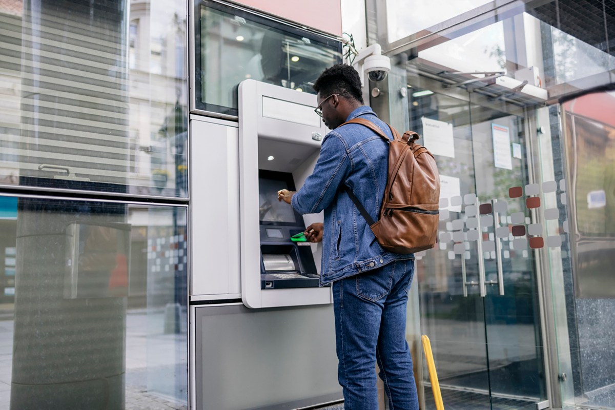 A person with a denim jacket and a backpack uses their credit card at an ATM to withdraw cash. The ATM is outside the bank entrance on the sidewalk of a city street.